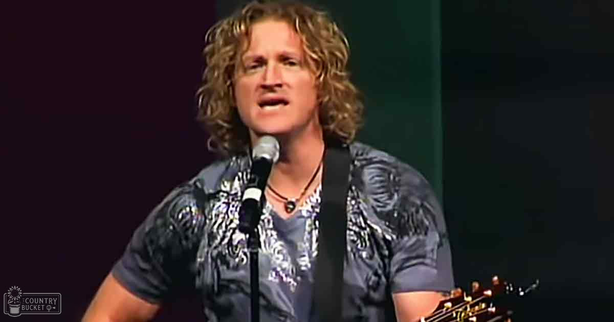 A middle-aged male singer wearing a floral patterned, short-sleeved shirt with rounded neckline and short sleeves, performing on stage at what appears to be a concert or music event.