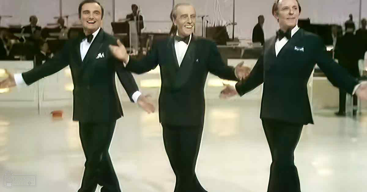 A group of adult male performers dressed in formal black tuxedos with white shirts and bow ties, set against an indoor studio or stage backdrop resembling ice skating rink flooring.