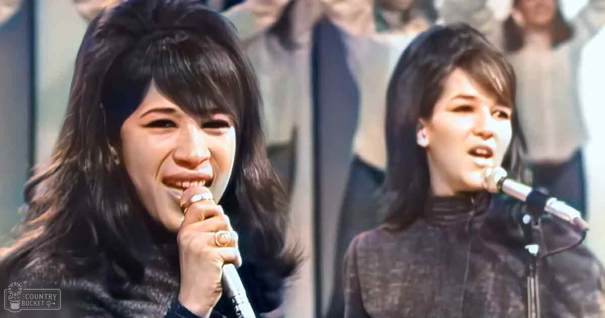 A young adult female singer wearing a black lace top at an outdoor event, with another person in a dark textured jacket visible in the background.