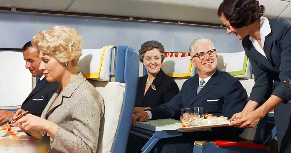 Interior view inside an airplane cabin from the mid-20th century; passengers in formal attire, flight attendant serving food, muted blue and white color palette with yellow trim on seats.