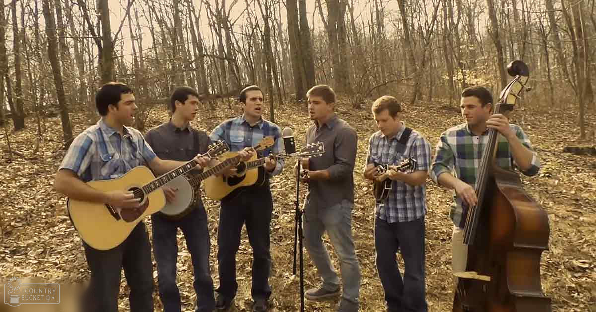 A group of young adults playing music outdoors in a forested area with bare trees and fallen leaves.
