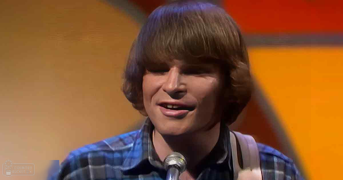 Young adult male in a relaxed casual blue and white checkered button-down plaid shirt with short sleeves on stage under warm orange and yellow lighting, likely from a performance or interview.