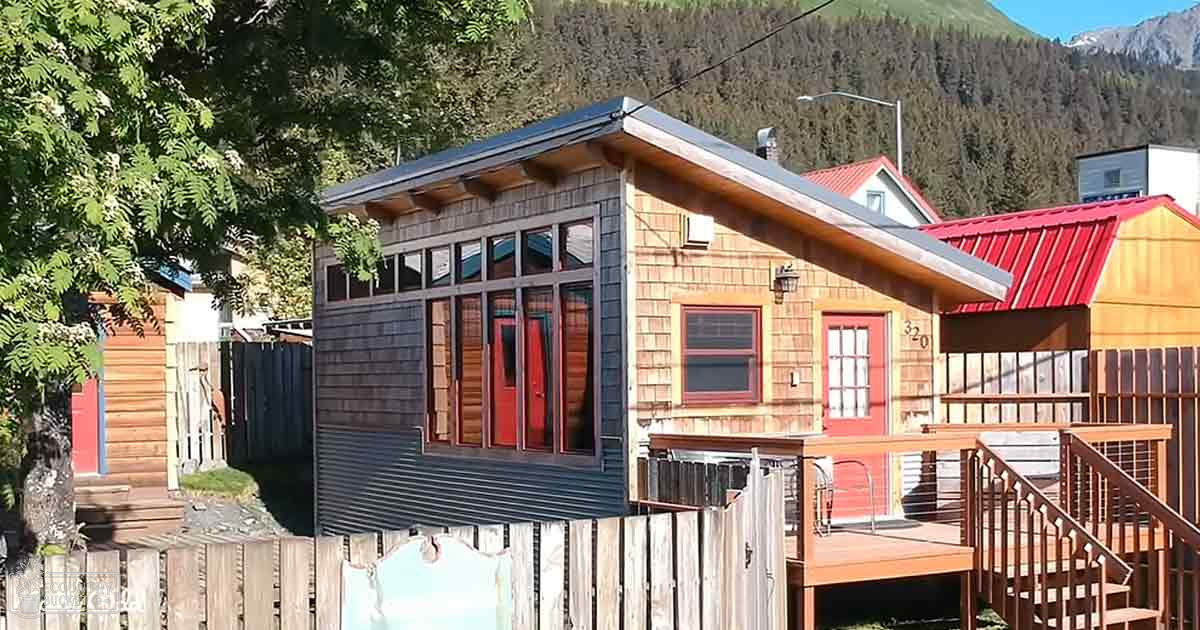 Small wooden cabins with red doors and windows in a residential area surrounded by trees and mountains. Residential neighborhood with natural surroundings, number '320' visible on the side of one cabin.