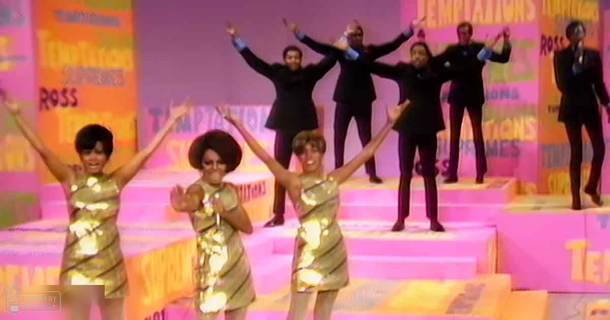 A performance scene with young adult female performers in gold sequined dresses, standing on a stage with pink and orange steps under well-lit daytime lighting.