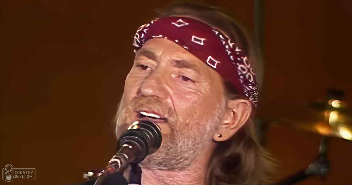 A middle-aged adult male singer wearing a red patterned bandana and small earring performs in an indoor music setting with musical instruments visible in the background, under warm lighting.