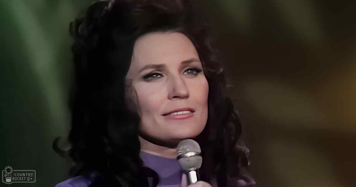A middle-aged woman in a purple, high-necked, long-sleeved garment from the 1960s or 1970s holds a microphone indoors with blurred greenery and plants in the background.