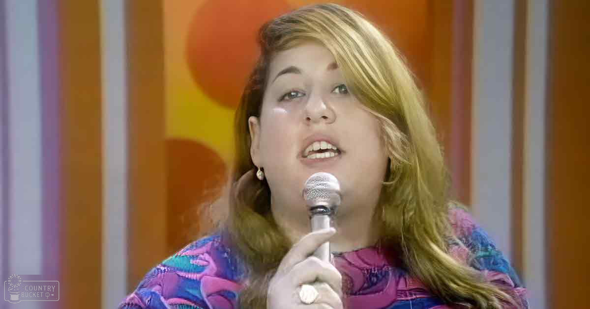 Young adult female wearing a bright pink, purple, and blue patterned long-sleeved top with pearl earrings; holding a microphone on stage with vertical striped background.