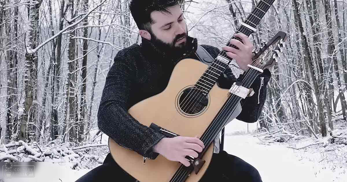 An adult male in a snowy forest, wearing a dark gray turtleneck sweater with long sleeves, playing a natural wood guitar with black accents.