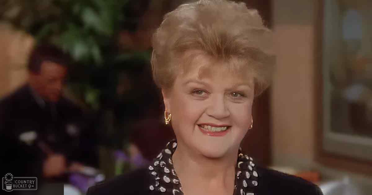 An older adult woman wearing a black top with white polka dots in an indoor social setting, surrounded by out-of-focus figures and greenery.