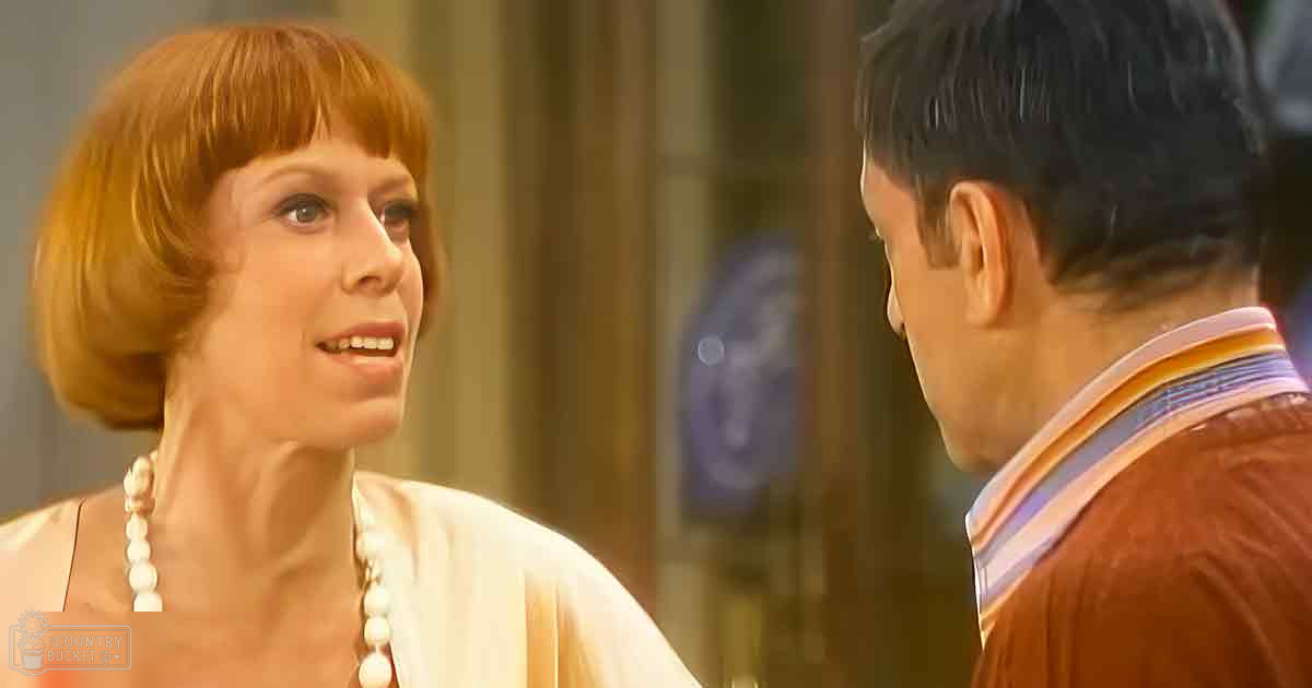 A middle-aged woman wearing a white blouse with high neckline and pearl accessories stands indoors near curtains and what appears to be part of a window or door frame, suggesting daytime lighting.