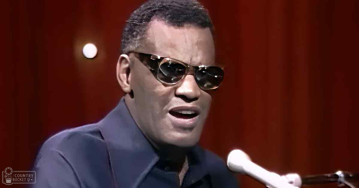 Middle-aged adult male singer wearing a tailored dark blue suit and aviator-style sunglasses, performing into a microphone against a red curtain backdrop.