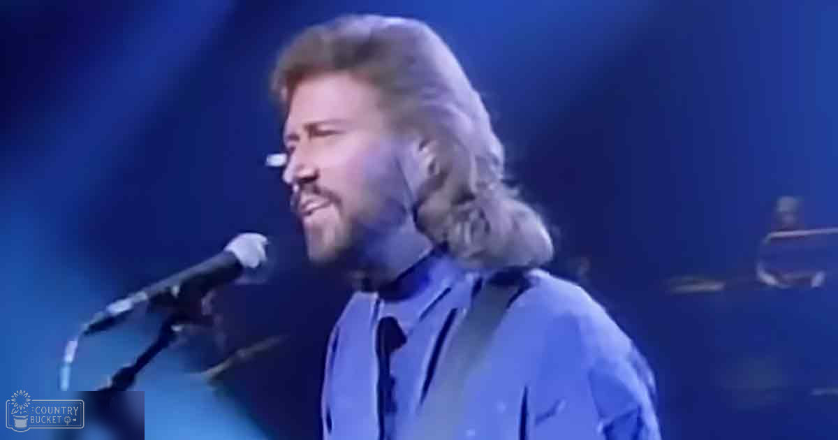 Middle-aged male singer in blue button-up shirt with tie, dark jacket or vest over it, performing into a microphone at what seems like a concert setting.
