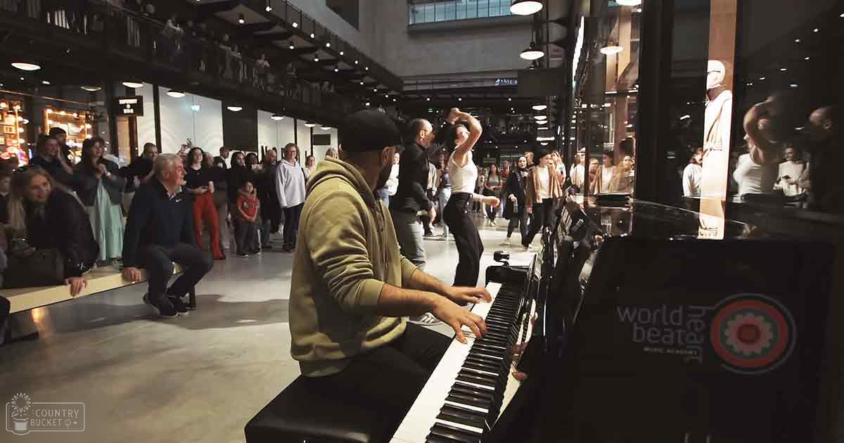 An adult male pianist in a beige hoodie playing at an indoor public space; audience includes middle-aged and older adults in casual attire, natural light from large windows suggests daytime.