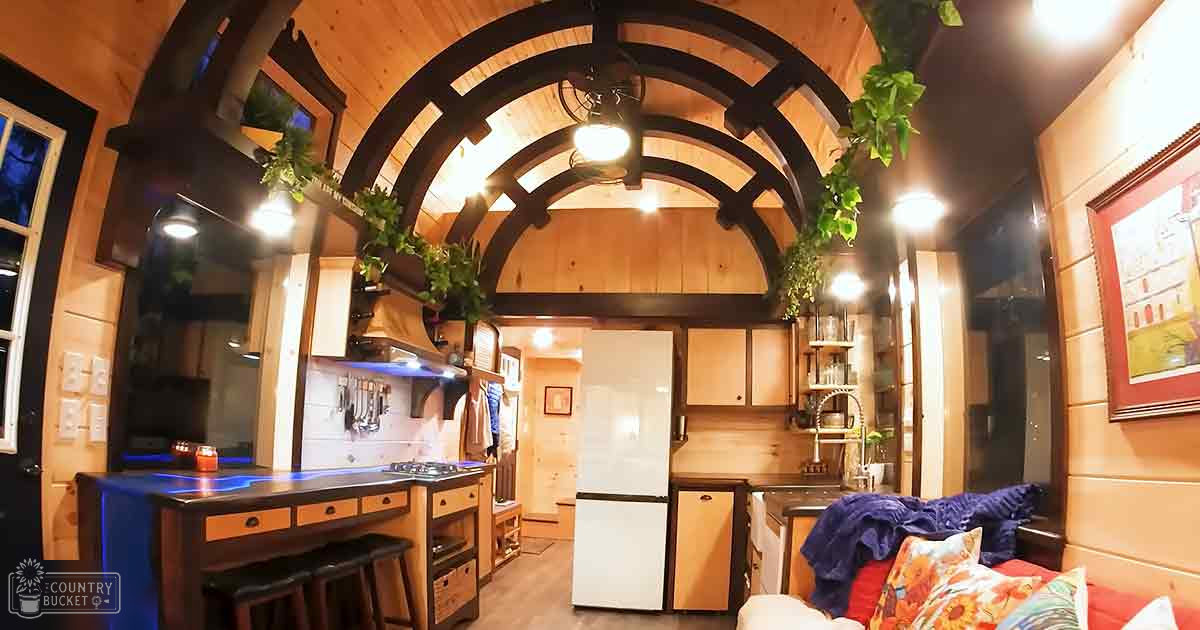 A cozy, compact cabin interior with wooden cabinets, a kitchenette, and seating area with warm tones like brown, beige, white, red/orange, and green from plants.