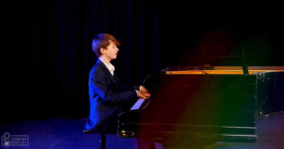 A young boy in a dark suit jacket plays a grand piano with glossy black finish, set against dark curtains suggesting an indoor performance venue.