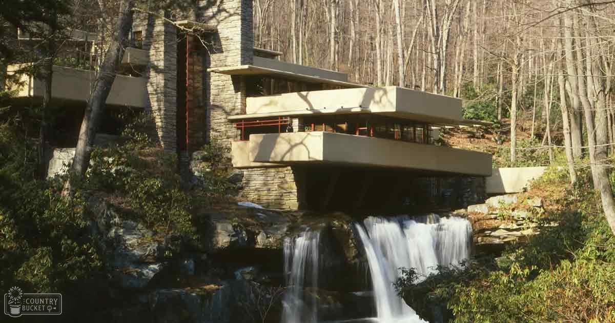 A modernist house with cantilevered concrete slabs over a waterfall in Pennsylvania, surrounded by late autumn or early spring trees and green vegetation on a sunny day.