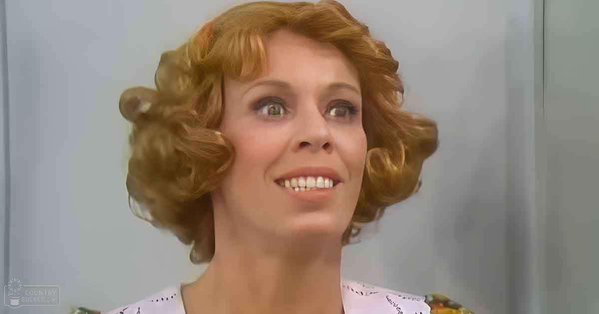 An adult female wearing a white floral-patterned blouse in an indoor setting with plain white walls and even bright lighting.