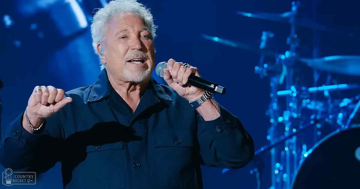 A middle-aged male singer wearing a dark blue button-up shirt performs on stage with a microphone, wristwatch, and bracelet under blue stage lighting.