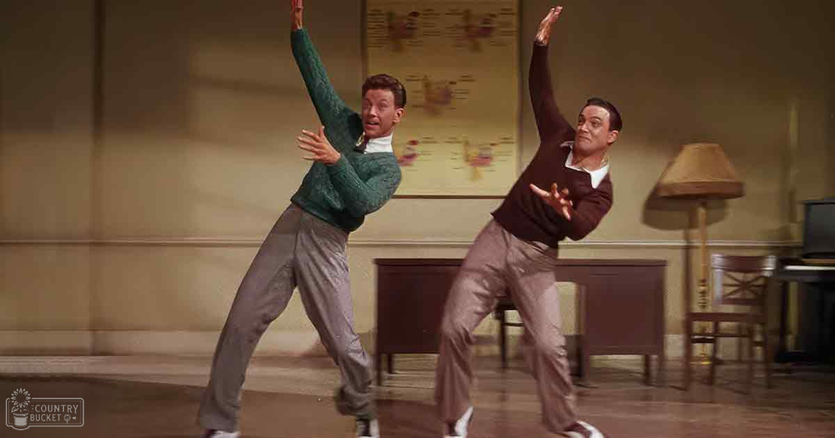 Adult male dancers in a dance studio setting with green and gray sweaters, beige lamp, artificial lighting.