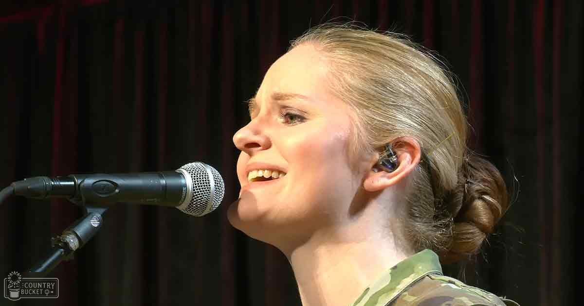 A young adult female in a camouflage-patterned shirt performs on stage with earbuds, set against dark curtains and indoor venue lighting.