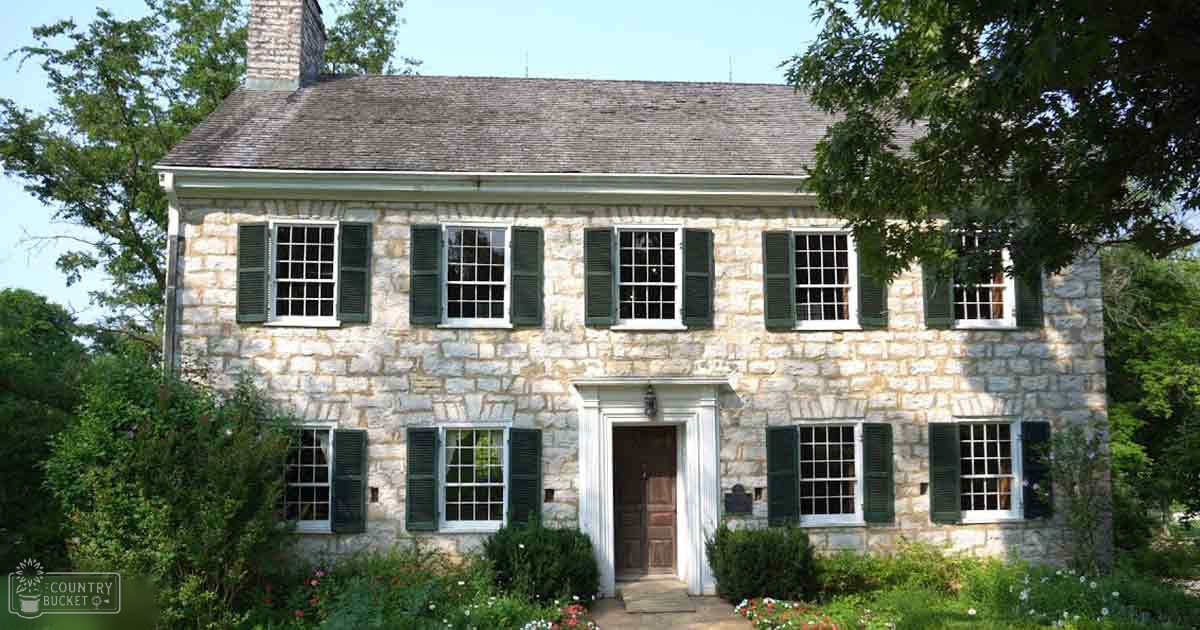 A historic stone house with multiple windows and shutters, surrounded by lush greenery under clear skies during midday.
