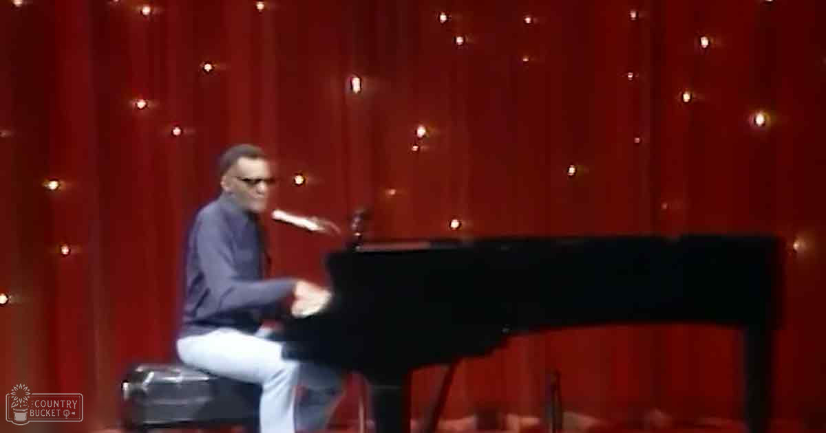 An adult male performer wearing a dark blue long-sleeve shirt and light-colored jeans plays piano in a stage setting with red curtains and fairy lights, suggesting a formal indoor performance space.