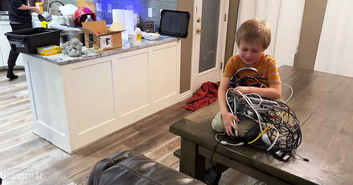A toddler wearing a striped orange and yellow t-shirt sits at a wooden table with tangled wires and electronic devices, amidst various kitchen items like Amazon boxes and cleaning supplies in an indoor kitchen setting.