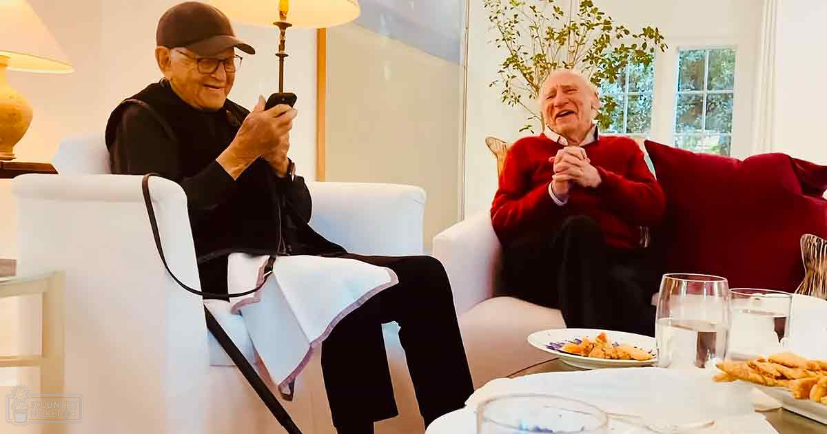 Two older adults seated in a living room with white furniture and greenery visible through the window, suggesting daytime in spring or summer.