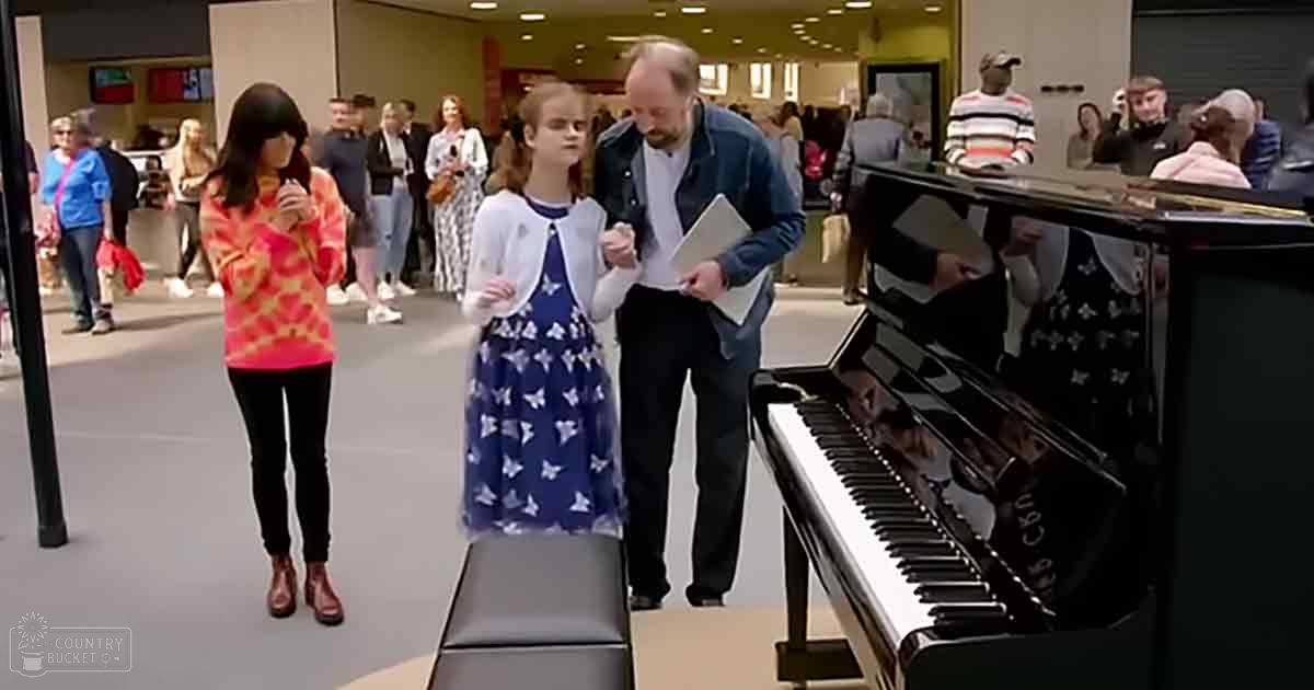 A young girl in a blue dress with white butterfly patterns stands before a grand piano as an adult holds a tablet or small book nearby. The scene takes place indoors, possibly at a public event like a shopping mall.
