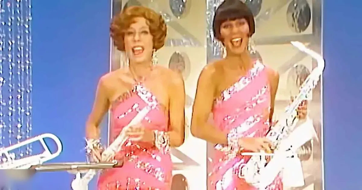 Two adult female performers wearing matching pink floral one-shoulder dresses, seated on a stage with a microphone stand and decorative backdrop.