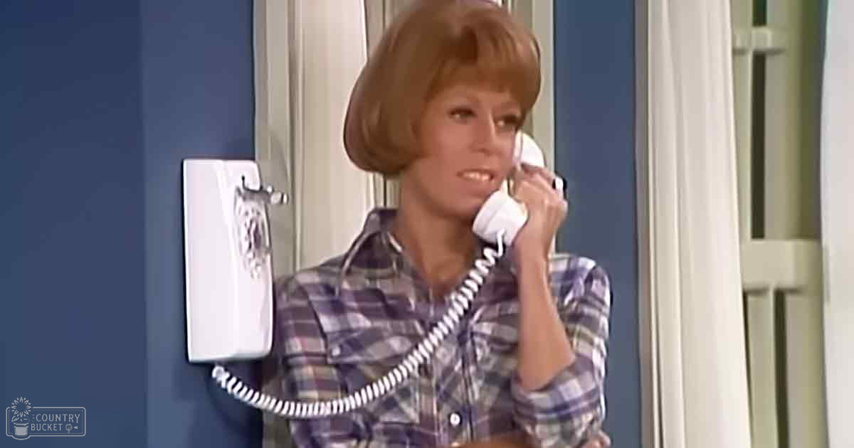 An adult female wearing a mid-20th century-style plaid button-down shirt indoors, holding a white corded phone receiver to her ear with even daytime lighting.