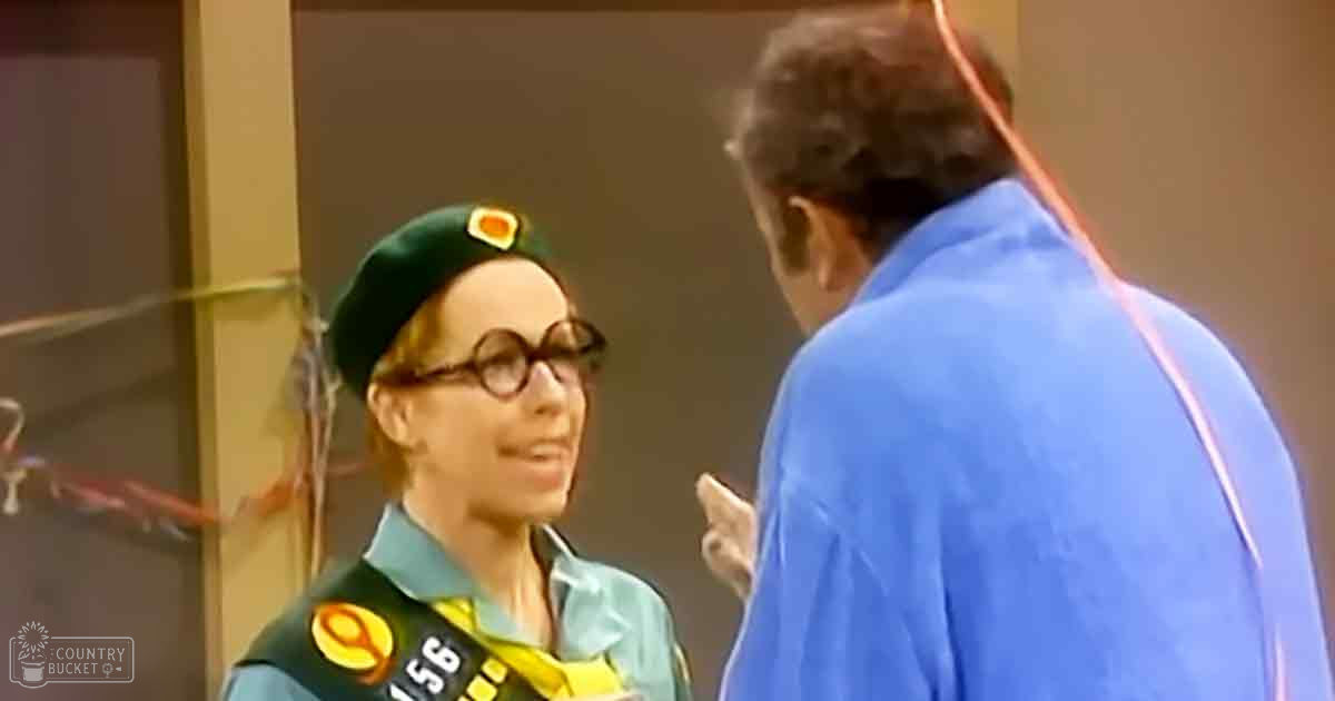 A young adult female scout in a green beret with an orange emblem, blue shirt, dark green vest with yellow and red patches stands indoors. An older male figure is present to her right.