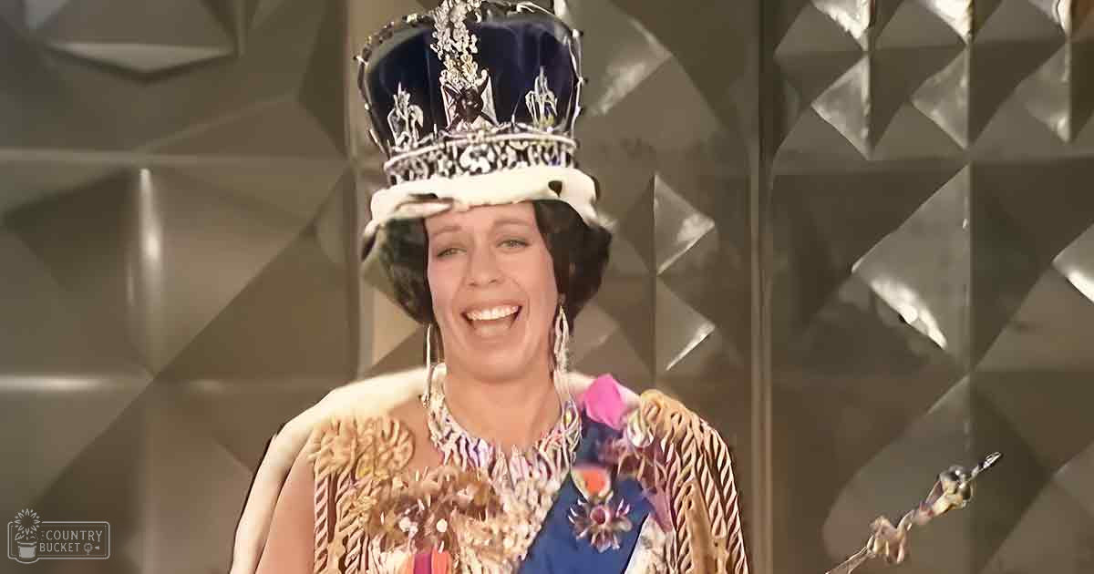 An adult female performer in a theatrical setting, wearing an ornate crown and a sleeveless blue floral-patterned gown with gold embroidery. She holds a scepter or staff.