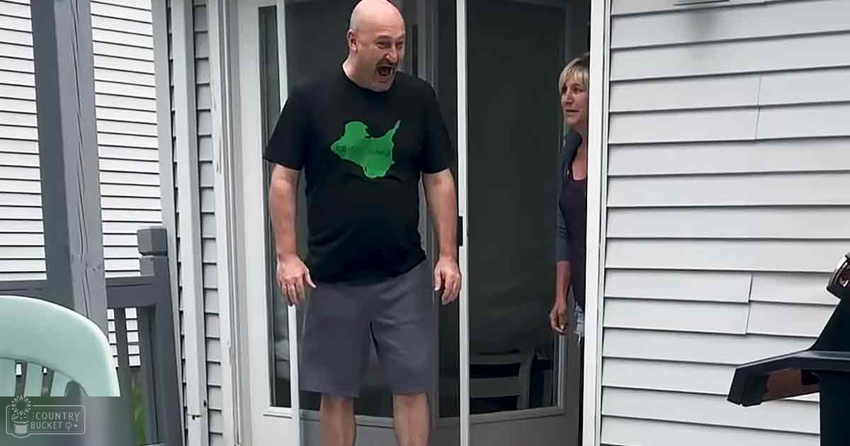 A middle-aged man in a black T-shirt with green graphic interacts near the entrance of a residential home, surrounded by outdoor furniture and musical equipment on a light gray vinyl siding porch.