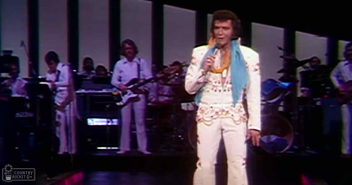 A middle-aged male performer in a white floral-patterned suit with a blue scarf, holding a microphone, stands center stage at what appears to be a concert or theatrical performance. Background includes other performers and musical equipment.