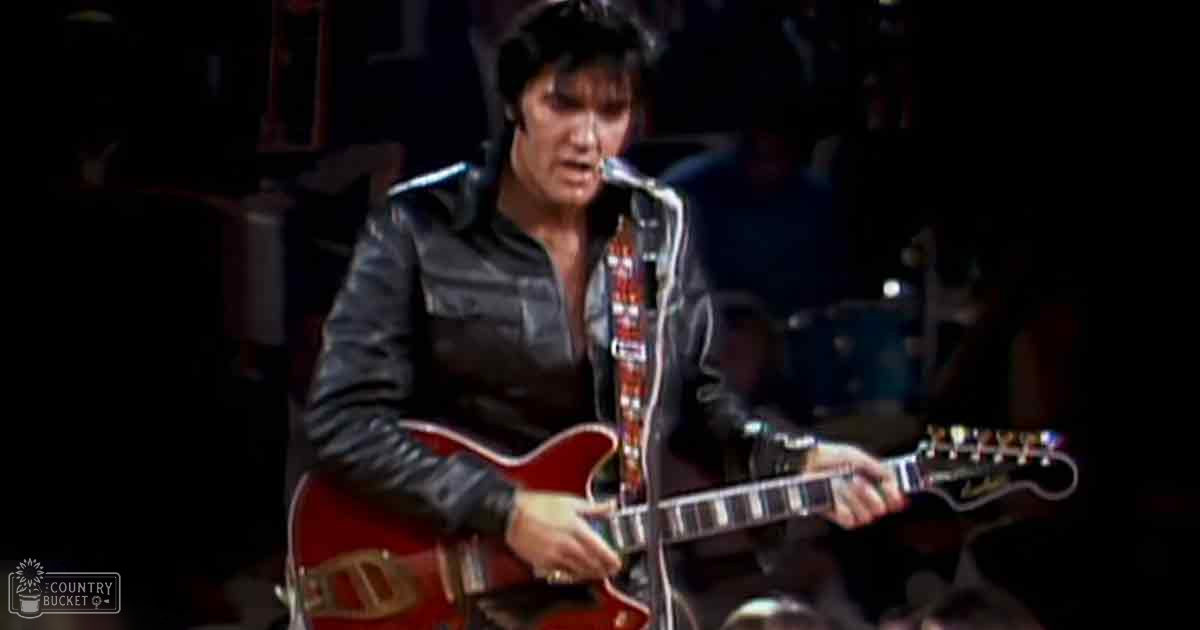 Adult male singer performing on an indoor stage with a microphone stand, wearing a form-fitting black leather jacket from the 1950s-1960s rock and roll era.