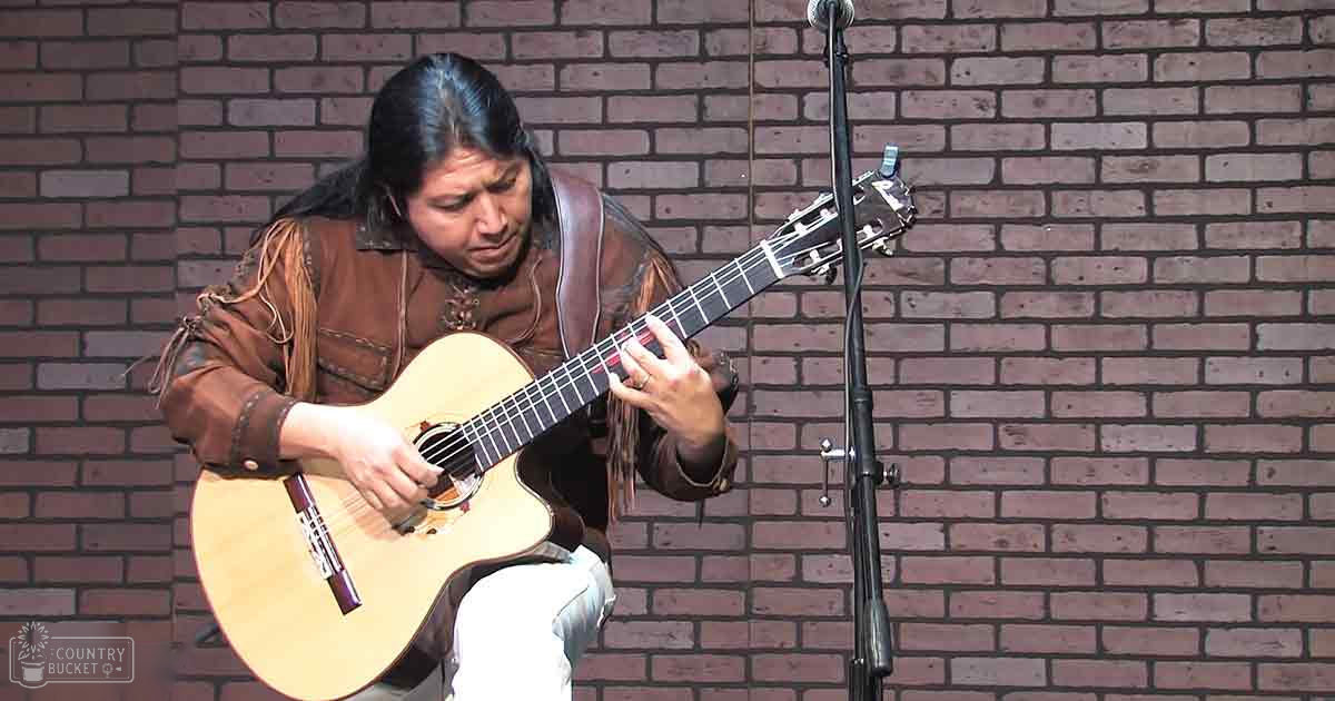 An adult female musician in a traditional outfit plays guitar against a brick wall backdrop.