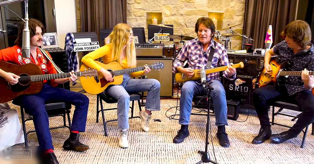 A group of musicians in a recording studio; adult male singer wearing red floral shirt, young female singer in yellow top, adult male musician in plaid shirt, and another young male musician in patterned dark shirt with acoustic guitars, electric guitar, bass guitar, amplifiers labeled 'Blues 35' and 'SR10', microphone stand, reserved sign for John Fogerty, chairs for musicians; music studio setting indoors with curtains and controlled lighting.