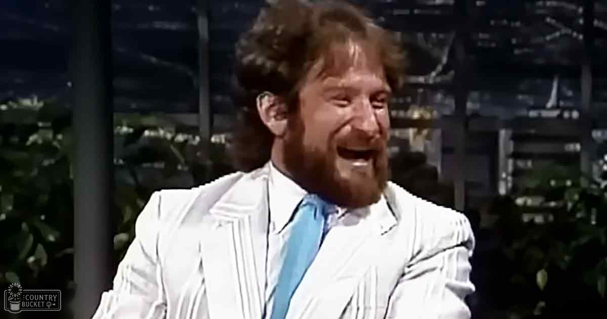 A middle-aged man with a beard and curly hair wearing a white 1970s-style suit jacket in an indoor office or studio setting, illuminated by artificial light.
