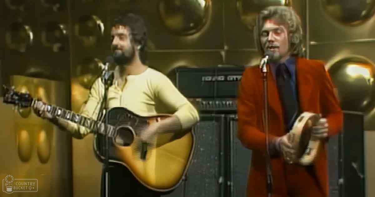 Musical performance by two adult males in an indoor venue, with one wearing a light-colored shirt and the other in a bright orange coat.
