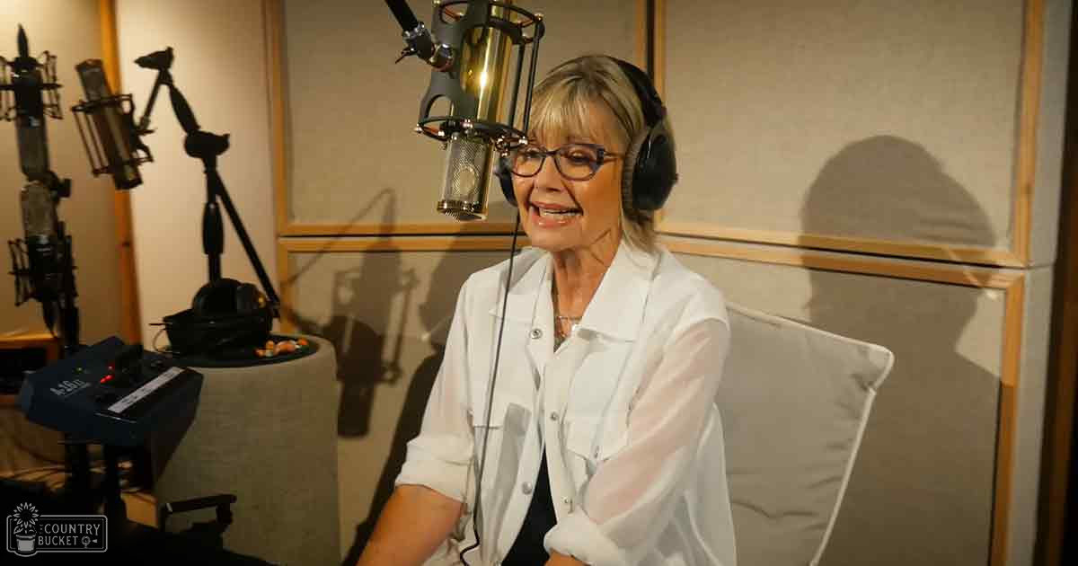 A middle-aged adult female in a recording studio wearing a white button-up shirt with long sleeves over a black top, surrounded by professional audio equipment.