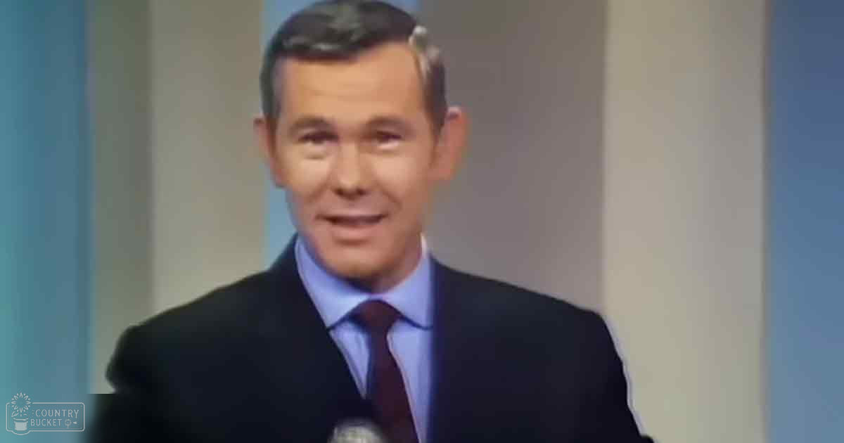 A middle-aged adult male in a professional setting, wearing a dark suit jacket with a light blue dress shirt and maroon tie. The background is plain with vertical lines, suggesting an indoor studio or office environment.
