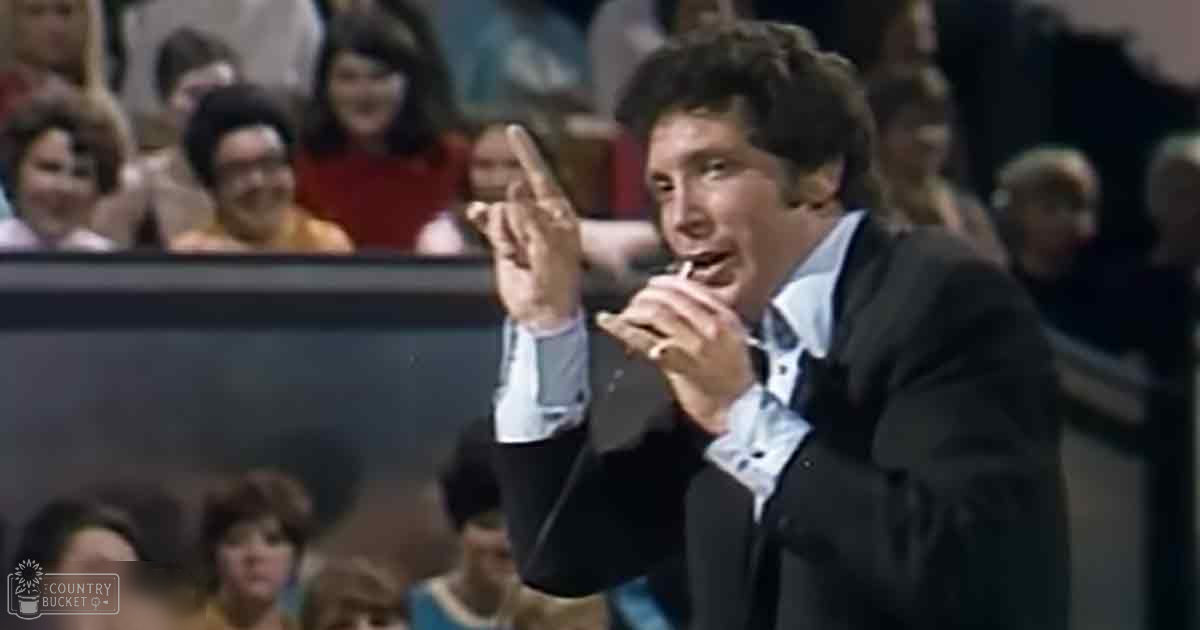 An adult male in a dark suit jacket over a light-colored shirt is gesturing at an indoor sports event with children and adults seated in rows, under artificial lighting.