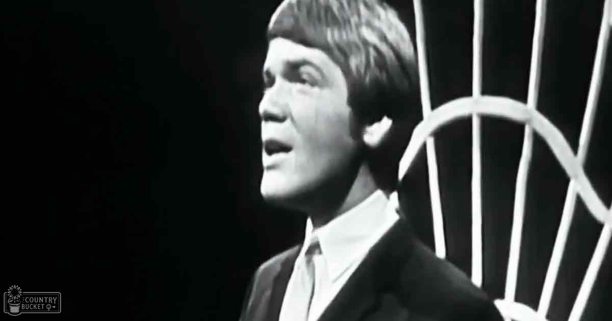 A young adult male in a dark suit jacket with a white collared shirt, standing indoors under dim lighting.