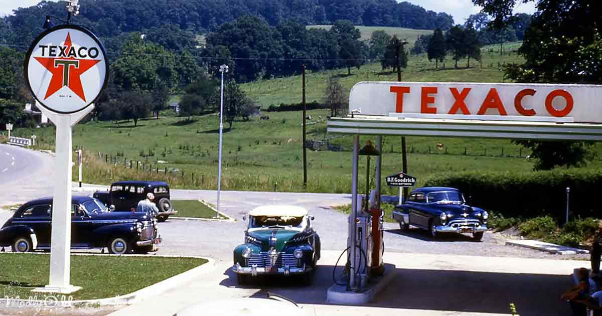 A vintage Texaco gas station in an open rural area with classic cars, lush greenery, and a clear sky during spring or summer.