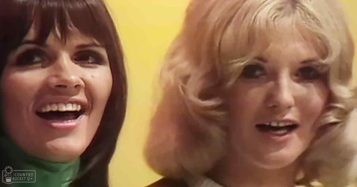 Two women in a studio setting wearing garments from the late 1960s to early 1970s; dark green turtleneck sweater and light-colored high-neck garment against a plain yellow backdrop.