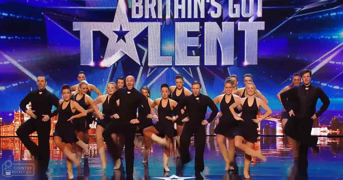 A dance group performing at 'Britain's Got Talent' with adults wearing black outfits, set against a large screen displaying the show logo and an urban cityscape.