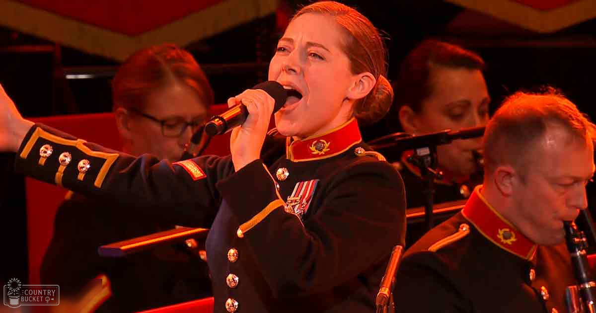 A young adult female singer in a military-style uniform performs into a microphone on an indoor stage with red lighting and musical instruments visible.