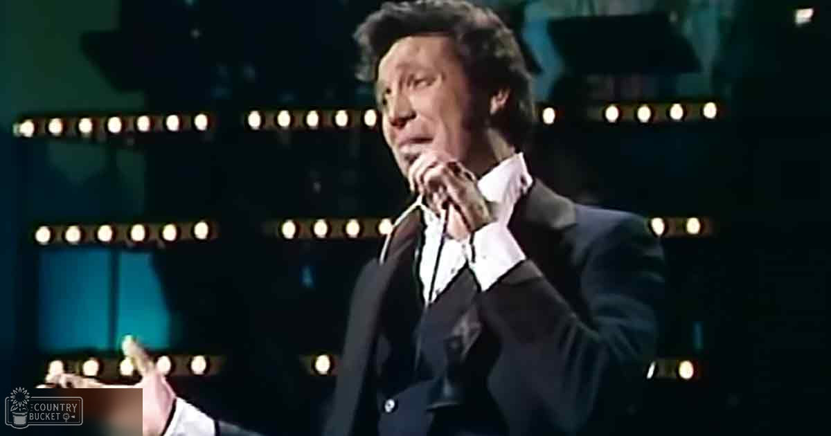 A middle-aged male performer in a dark suit jacket with 'Madly Odd' printed on his garment, set against bright stage lights during an indoor performance.