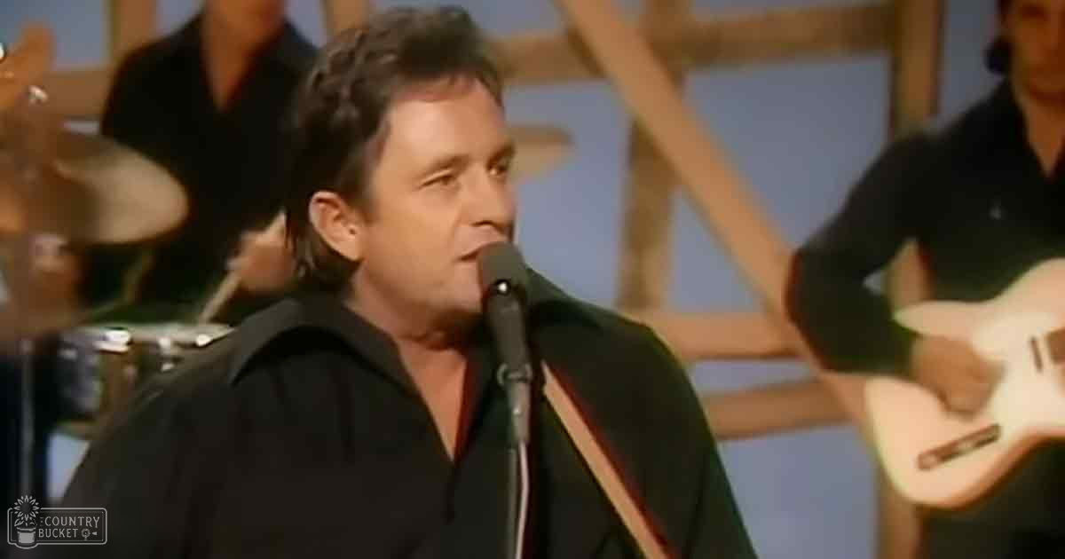 A middle-aged man singing into a microphone, wearing a black jacket and white shirt, with two young adults playing drums and holding guitars in a studio setting.